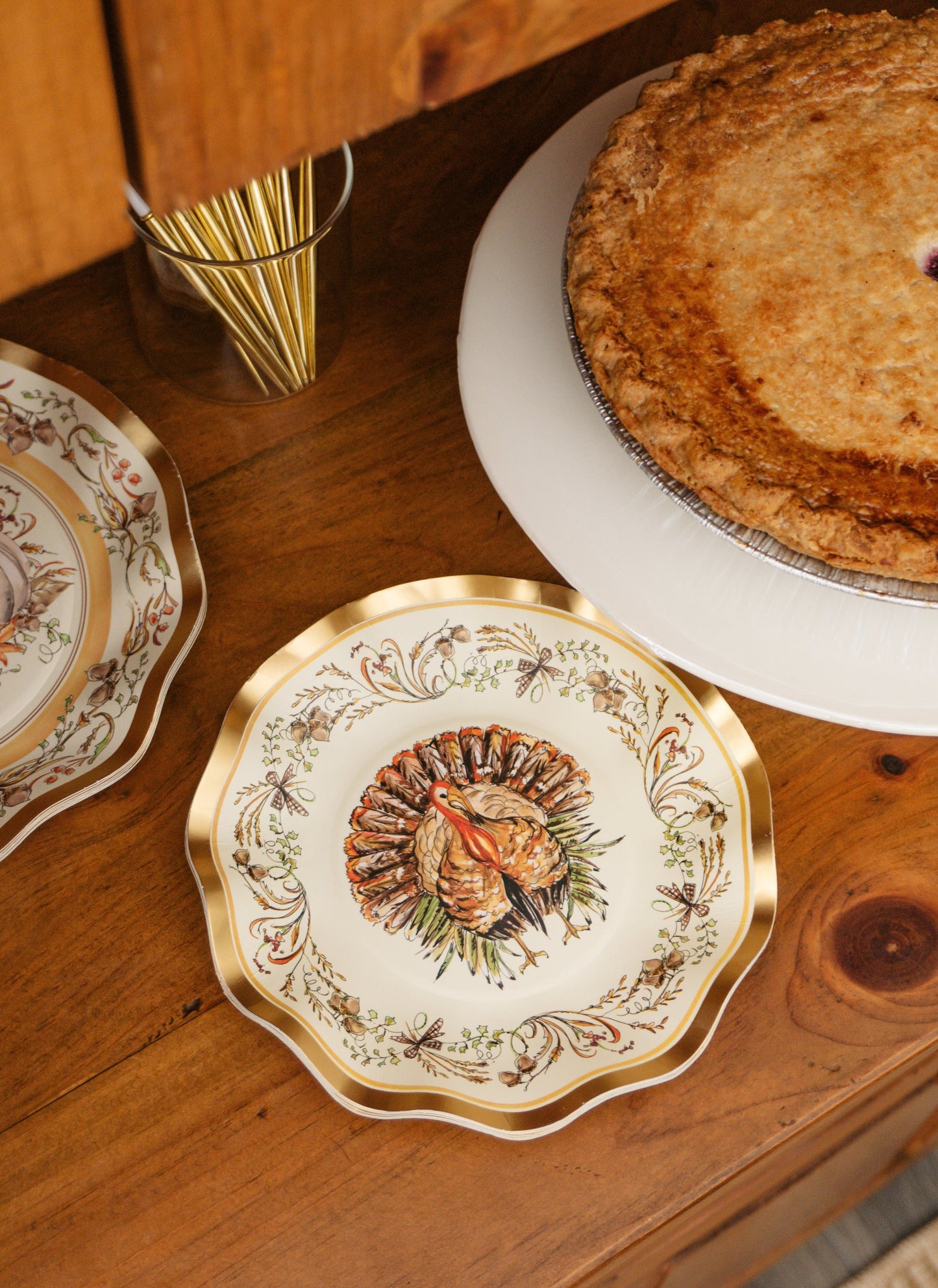 A decorative Turkey & Gourds: Thanksgiving Table for 16 setting featuring a turkey illustration on a disposable party plate, placed on a wooden surface next to a whole pie and a glass filled with gold cutlery.
