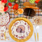 Create a festive Thanksgiving tablescape with the Amber Harvest Table Setting by Sophistiplate, featuring turkey-themed designer disposable plates, gold chargers, floral napkins, gold utensils, and autumn blooms atop a plaid tablecloth. The elegant Amber Harvest collection adds sophistication to your celebration, with a charming honeycomb turkey decoration completing the look.