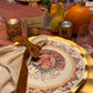 A Turkey & Gourds: Thanksgiving Table for 16 setting featuring a heavy duty paper plate with a turkey illustration, a beige napkin tied with a brown ribbon, and a place card labeled Nandinee, surrounded by gourds and candles on a red tablecloth.