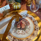 A Sophistiplate LLC Turkey & Gourds: Thanksgiving Table for 16 setting featuring a white turkey-illustrated heavy duty paper plate layered over a gold charger, complete with a cloth napkin, velvet ribbon tie, and a place card labeled Phil.
