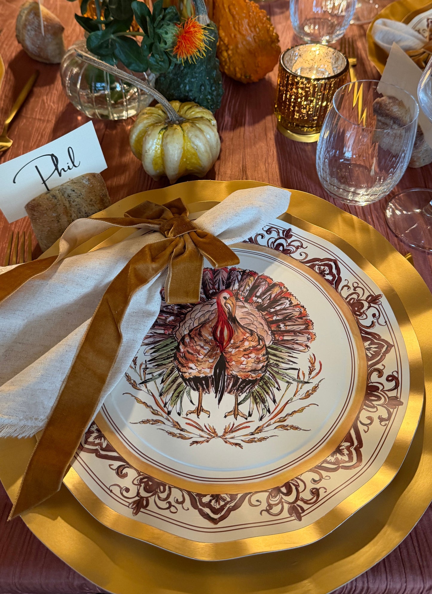 A Sophistiplate LLC Turkey & Gourds: Thanksgiving Table for 16 setting featuring a white turkey-illustrated heavy duty paper plate layered over a gold charger, complete with a cloth napkin, velvet ribbon tie, and a place card labeled Phil.