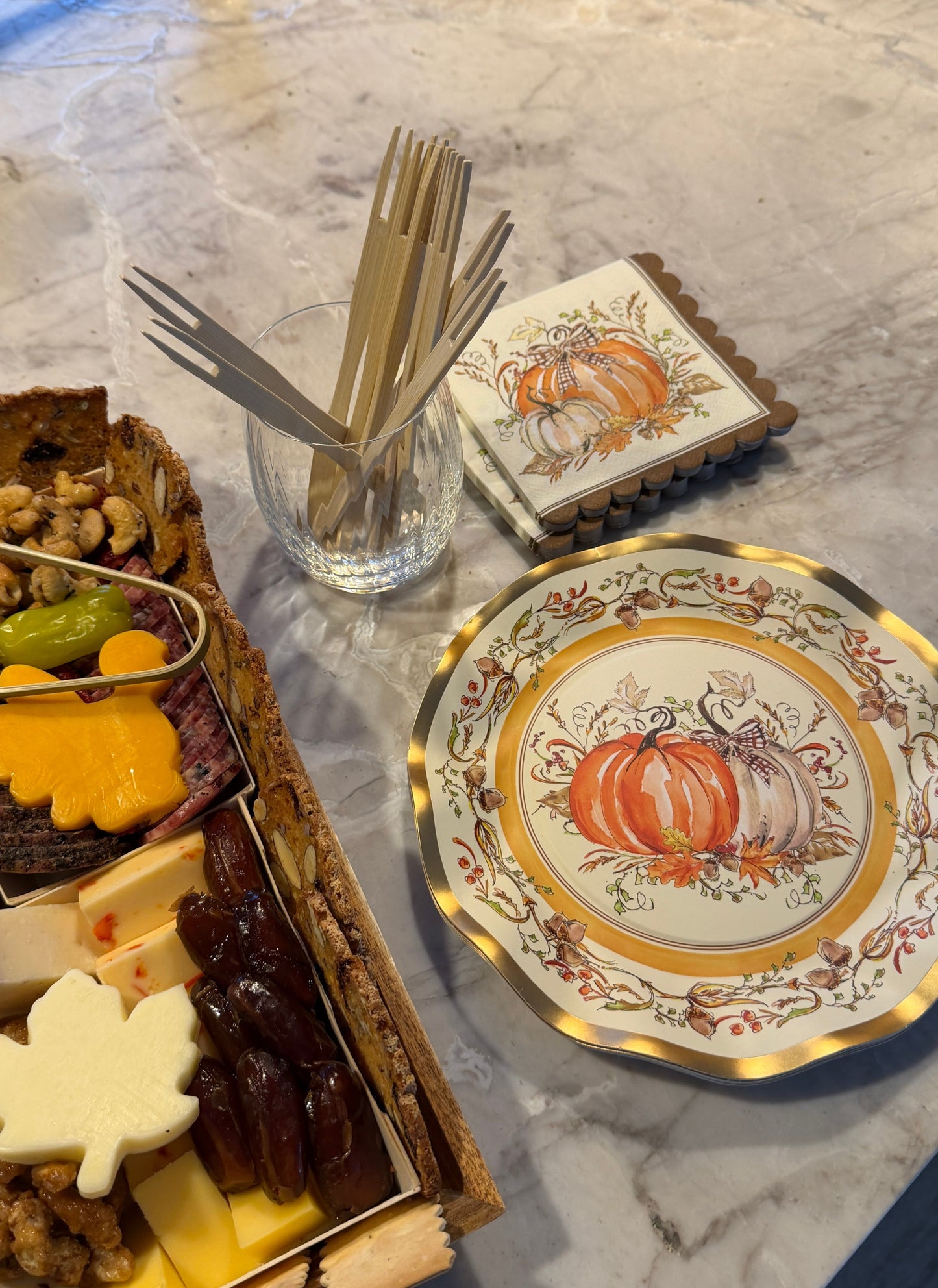 A charcuterie board next to Turkey & Gourds: Thanksgiving Table for 16 decorative plates and pumpkin-patterned napkins, with a glass holding wooden appetizer forks.