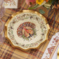 The Sophistiplate LLC Turkey & Gourds Table Setting Bundle features a decorative plate with a detailed turkey illustration at the center and a floral border, elegantly arranged on a plaid tablecloth alongside glassware and cutlery—a perfect example of designer disposable plates that blend festive style with convenience for any holiday gathering.