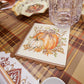 The Turkey & Gourds Table Setting Bundle by Sophistiplate LLC features festive napkins adorned with pumpkin illustrations and a bow, arranged on a plaid tablecloth alongside stylish disposable plates, a clear glass, and autumn-inspired tableware, creating a cozy Thanksgiving or fall-themed atmosphere.