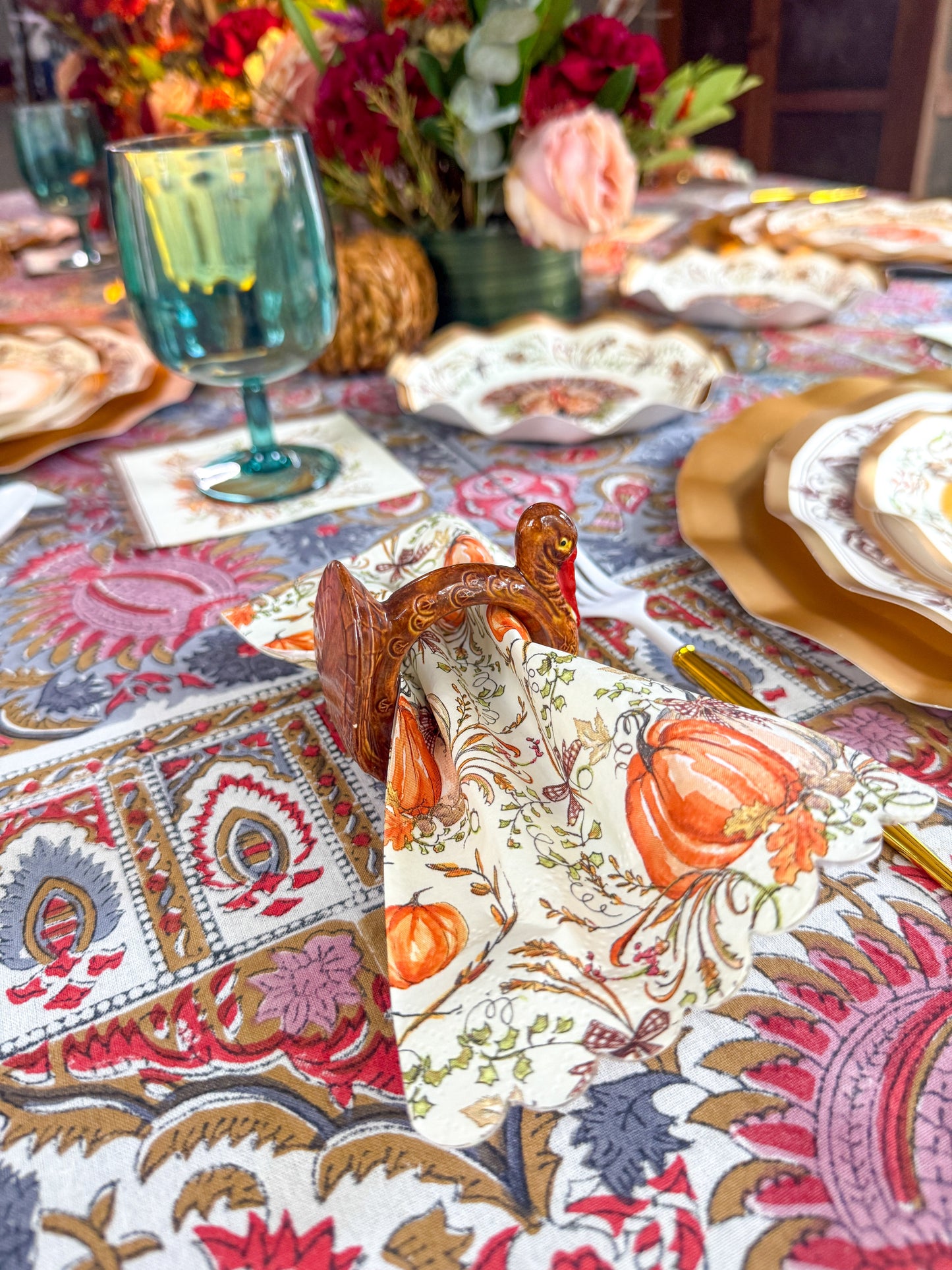 A Turkey & Gourds Guest Towels/20pk harvest pattern napkin decorated with pumpkins and autumn leaves, tucked into a turkey-shaped napkin ring on a patterned tablecloth.