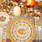 Create a warm and inviting Thanksgiving tablescape with the Turkey & Gourds Table Setting by Sophistiplate, featuring pumpkin-themed designer paper plates, gold chargers, faux pumpkins, candles, a floral garland, and vintage turkey napkins. White and gold flatware is arranged beside each place setting atop an orange tablecloth for a festive autumn look.