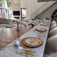 A Turkey & Gourds: Thanksgiving Table for 16 set on a long marble table with pumpkin-illustrated paper plates, gold chargers, patterned napkins, white cutlery, and wine glasses.